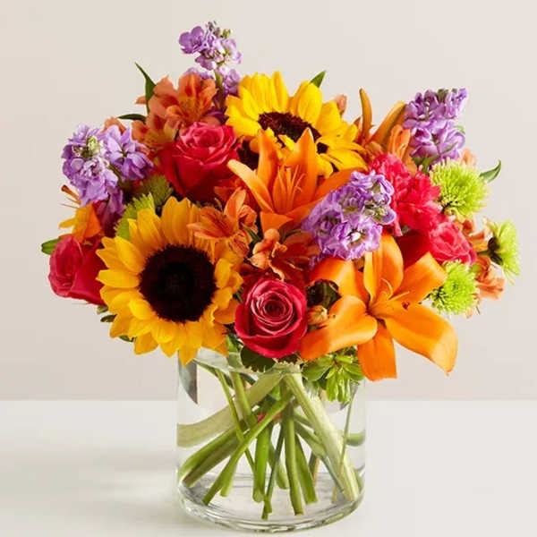 Bouquet of sunflowers, roses, lilies, and purple blooms in a glass vase