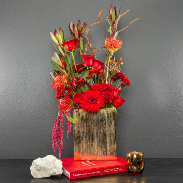 Red floral arrangement in a gold rectangular vase with a small gold candle holder.