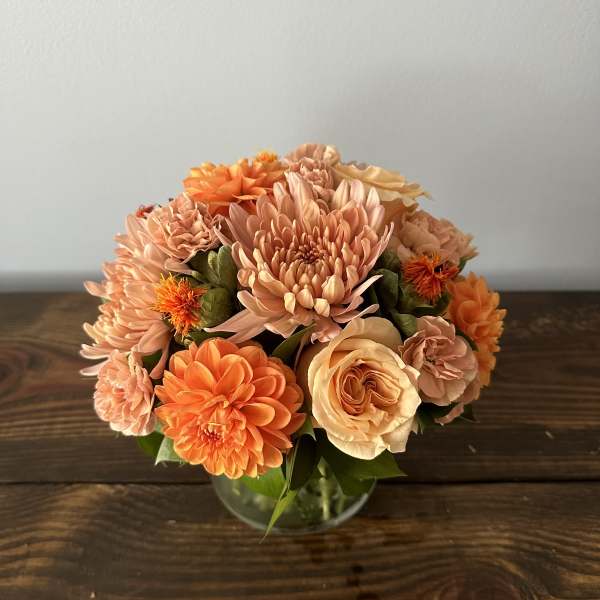 Short arrangement of orange and peach blooms in a clear glass bowl vase