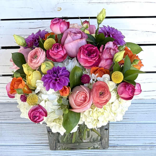 Colorful arrangement of pink roses, ranunculus, dahlias and white hydrangeas in a clear glass cube vase.