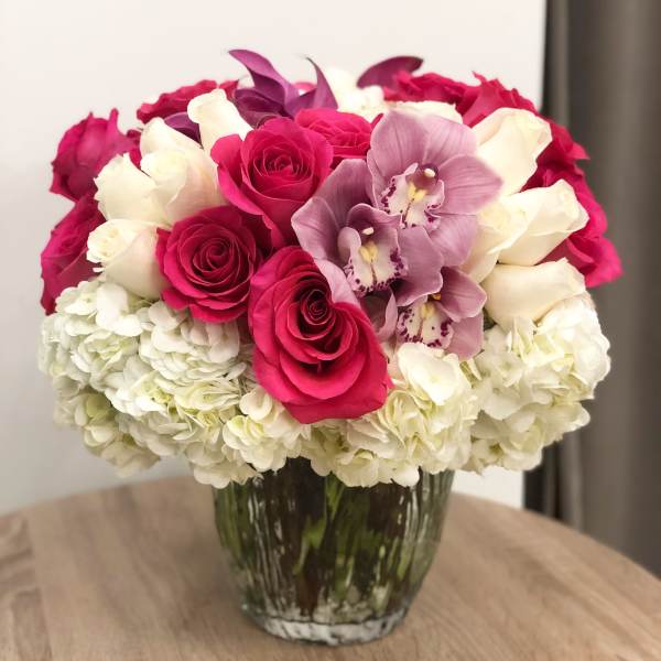 Round arrangement of hot pink roses, white roses, orchids, and hydrangeas in a clear glass vase.