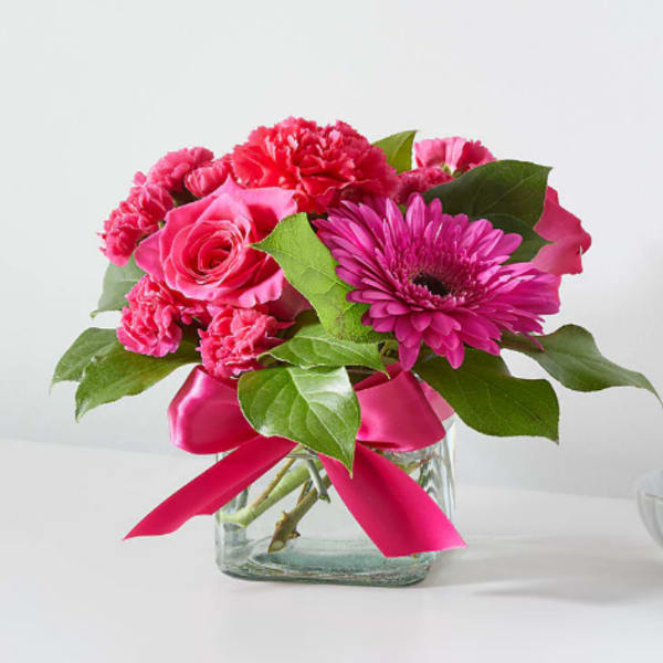 Bright pink roses, carnations, and a gerbera daisy in a square glass vase with a pink ribbon bow.