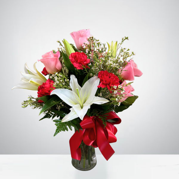Bouquet of pink roses, white lilies, and red carnations in a glass vase with a red ribbon