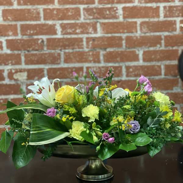 Low floral centerpiece with yellow roses, white lilies, and pink blooms in a pedestal bowl.