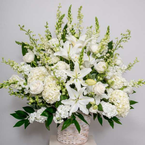 Large all-white arrangement of lilies, roses, hydrangeas, and snapdragons in a white woven basket.