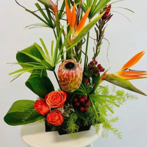 Tropical arrangement with orange birds of paradise, protea, and roses in a low black container