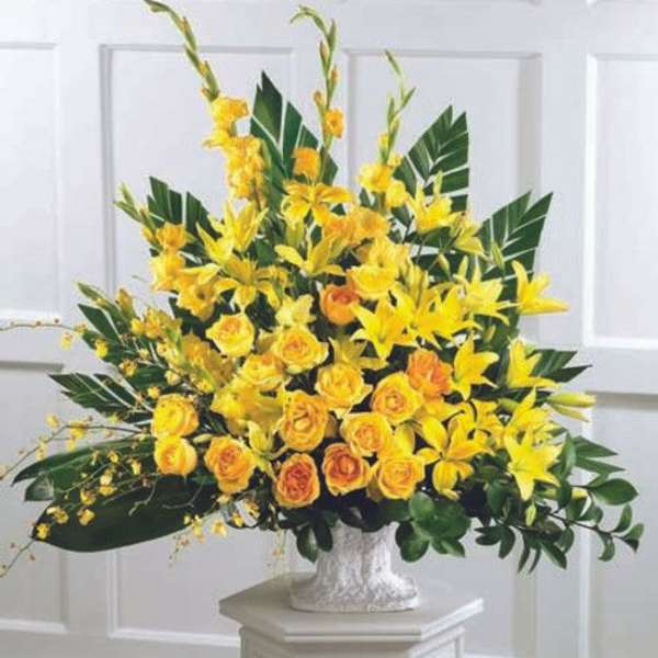 Tall yellow lily and rose arrangement with large green leaves in a white urn