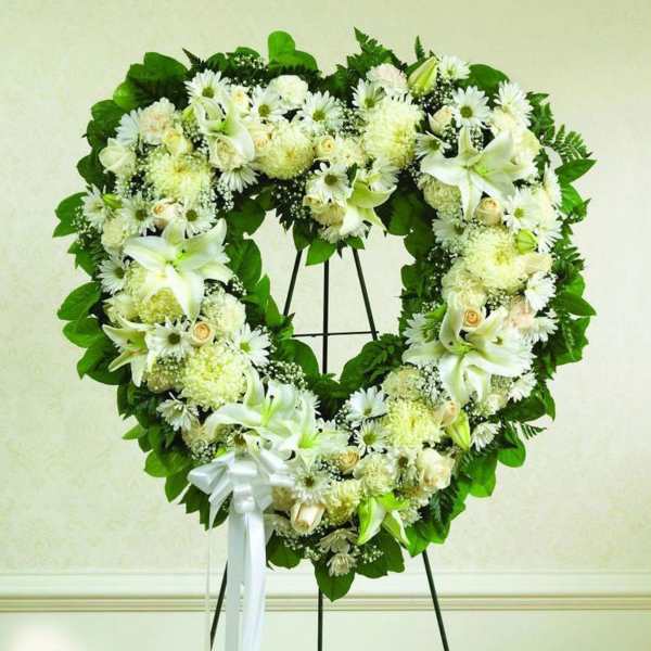 Heart-shaped standing wreath of white lilies, roses, and mums with green foliage and a white ribbon bow.
