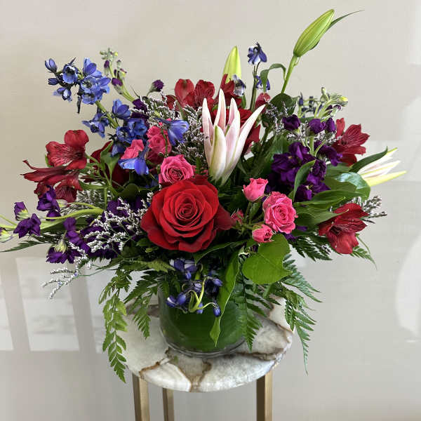 Colorful mixed arrangement with red roses, pink lilies, and purple blooms in a clear glass vase