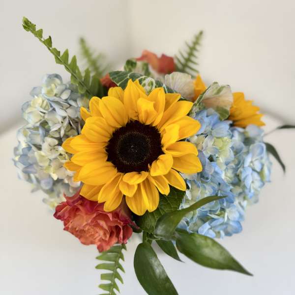 Compact bouquet with a large yellow sunflower, blue hydrangeas, and coral roses.