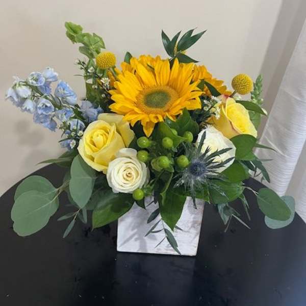 Bright sunflower and yellow roses with blue accents in a white cube vase
