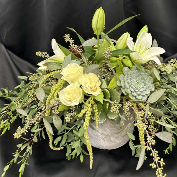 White lilies and pale roses in a low arrangement with trailing greenery and a succulent.