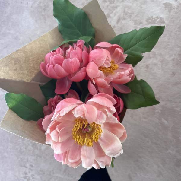 Small bouquet of pink peonies with yellow centers wrapped in kraft paper, held against a light background