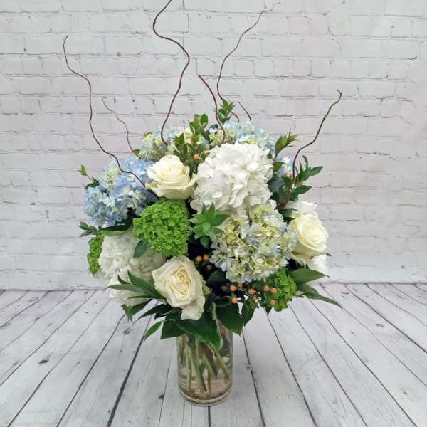 Tall glass vase arrangement with white roses, blue and white hydrangeas, green blooms, and curly willow branches.
