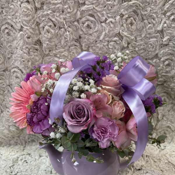Pink and purple flower arrangement in a lavender teapot vase with ribbon