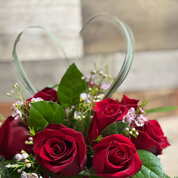 Short arrangement of red roses with small pink blossoms in a red glass cube vase with a heart-shaped grass accent.