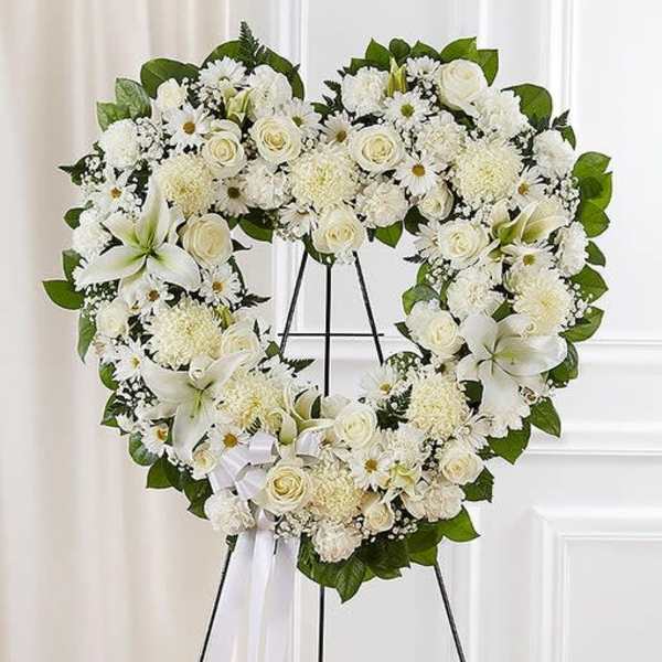 Heart-shaped white floral wreath with roses, lilies, and daisies on a stand