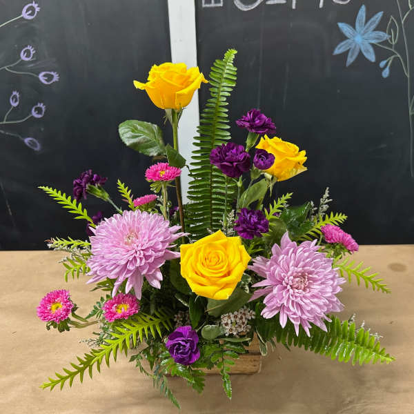 Mixed bouquet with yellow roses, lavender chrysanthemums, and purple blooms in a wooden container