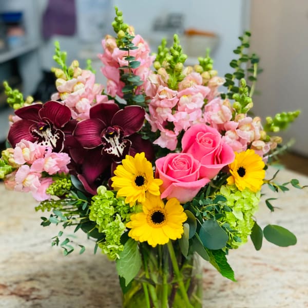 Mixed bouquet with pink roses, yellow daisies, and dark orchids in a glass vase
