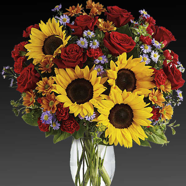 Bouquet of sunflowers, red roses, and small purple flowers in a clear vase