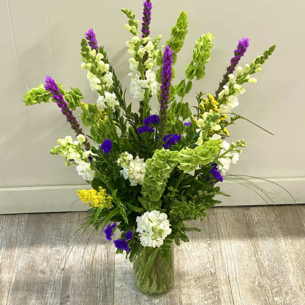 Tall mixed flower arrangement in a glass vase with purple, white, yellow, and green blooms