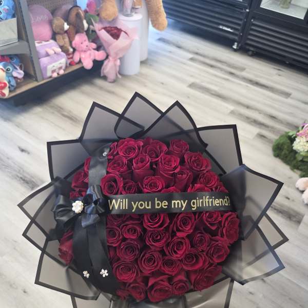 Bouquet of deep red roses wrapped in black paper with a "Will you be my girlfriend?" ribbon