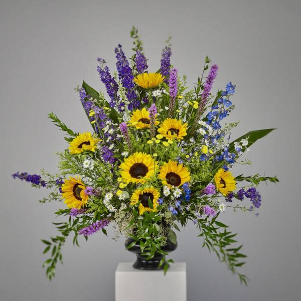 Tall arrangement of yellow sunflowers with purple and blue flowers in a black urn vase