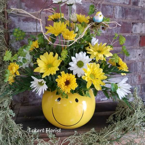 Yellow and white daisy arrangement in a smiley-face pot with a small bird decoration