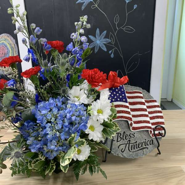Patriotic floral arrangement with blue hydrangeas, red carnations, and white daisies beside a flag plaque