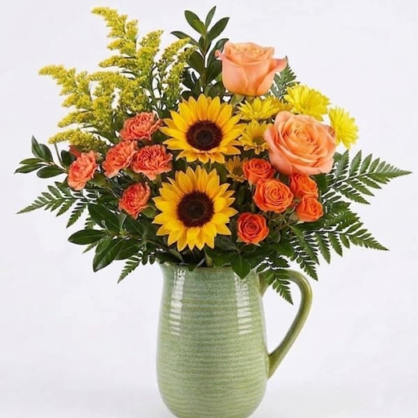 Bouquet of sunflowers, peach roses, and orange carnations in a green ceramic pitcher
