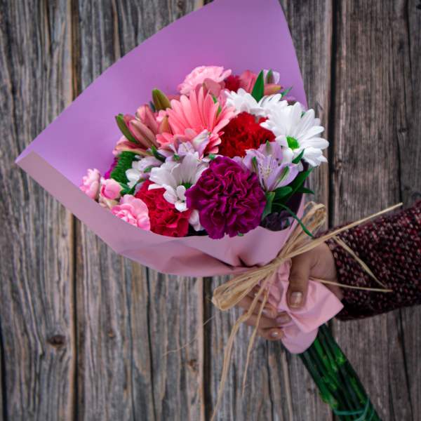 Handheld bouquet of pink, white, and red flowers wrapped in lavender paper