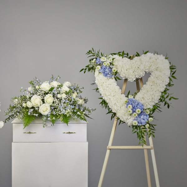 Sympathy set with white and blue arrangements and a heart-shaped wreath beside a white casket.