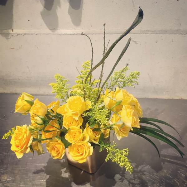 Yellow roses arranged in a square vase with tall greenery accents.