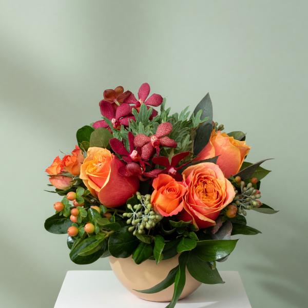 Orange roses and burgundy orchids arranged in a beige bowl