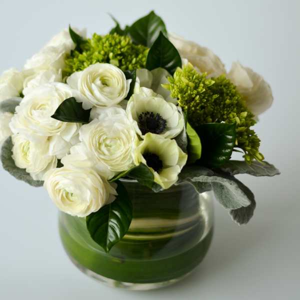 Low arrangement of white ranunculus and anemones with green hydrangea in a clear glass cylinder vase