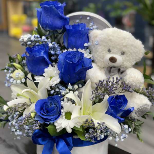 Blue roses and white lilies arranged with a teddy bear in a round box