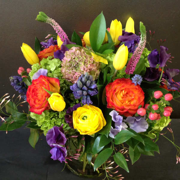 Bright mixed arrangement of yellow tulips, orange roses and purple blooms in a clear glass vase