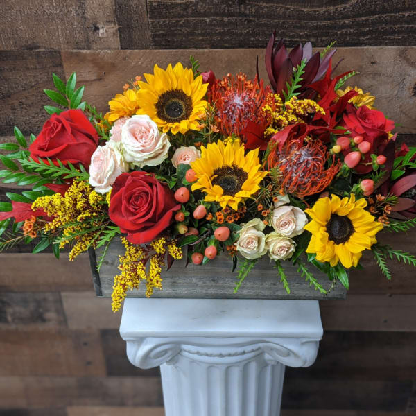 Arrangement of red roses, sunflowers, and orange tropical blooms in a rectangular container