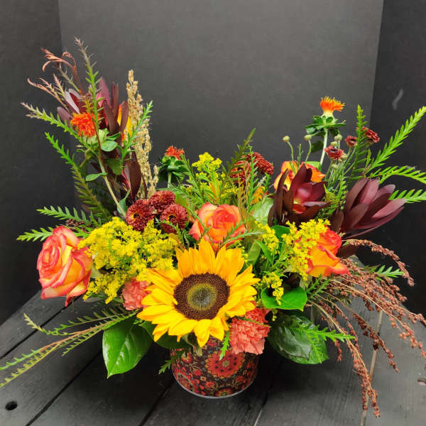Mixed bouquet with a sunflower, roses, and orange blooms in a patterned container