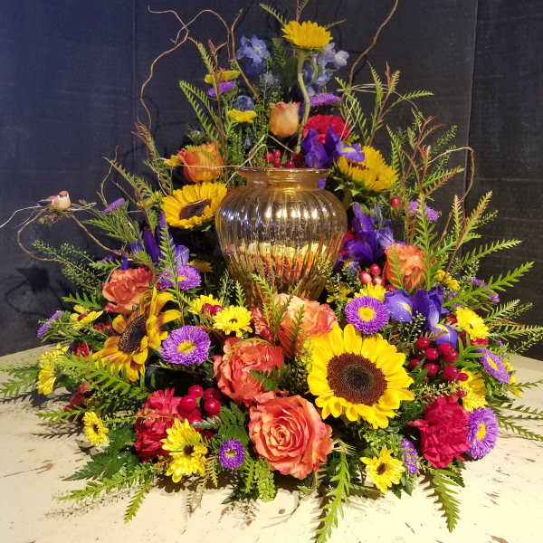 Large mixed floral arrangement with sunflowers, roses, and purple blooms in a gold vase