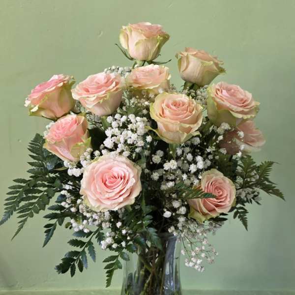 Tall arrangement of light pink roses and white filler flowers in a clear glass vase on a small dark table.