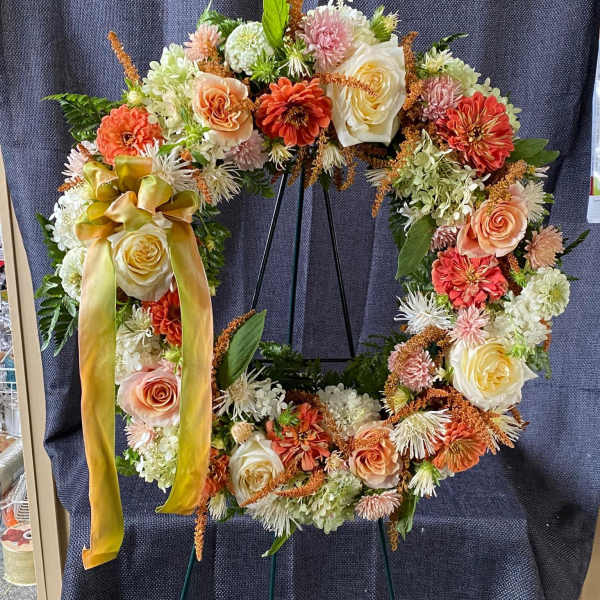 Floral wreath with peach, cream, and orange blooms on a stand with a ribbon bow