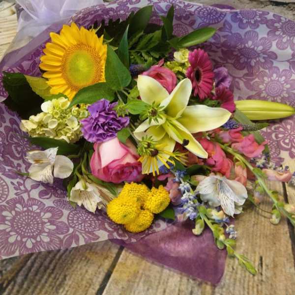 Mixed bouquet with a sunflower, lilies, roses, and purple blooms wrapped in purple paper