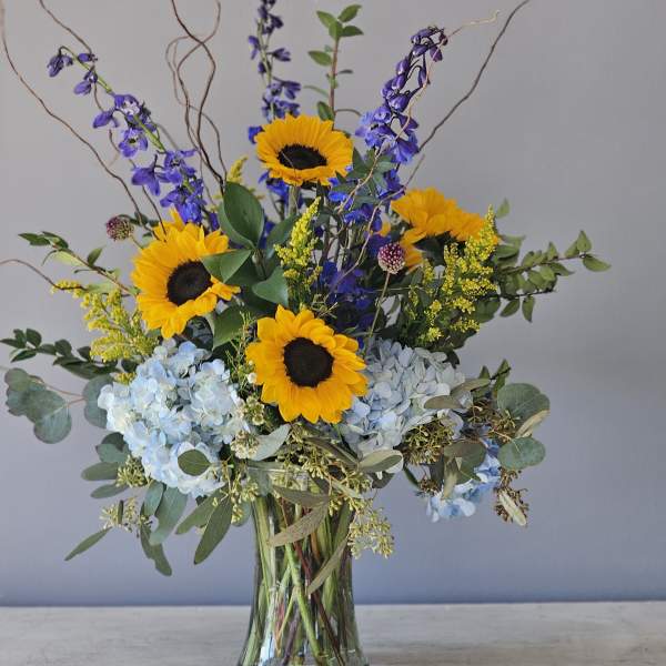 Tall arrangement of yellow sunflowers, blue hydrangeas, and delphinium in a clear glass vase
