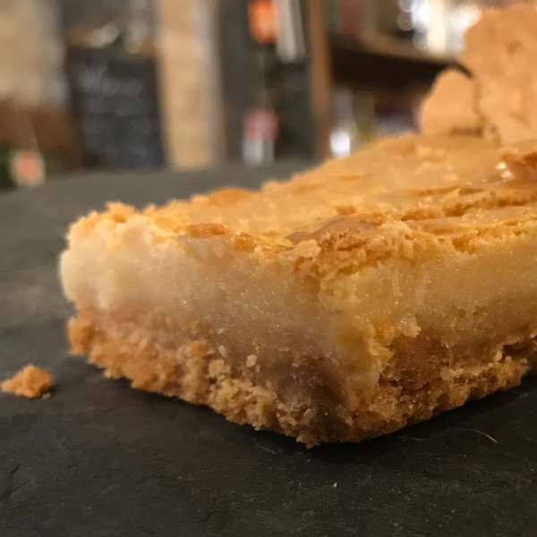 A square dessert bar with a crumbly golden crust on a dark surface
