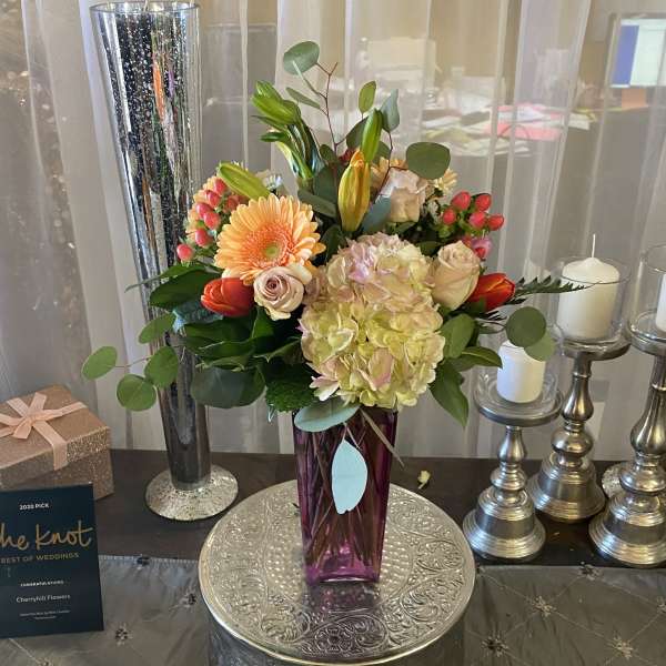 Mixed bouquet in a purple vase with candles and a gift box nearby