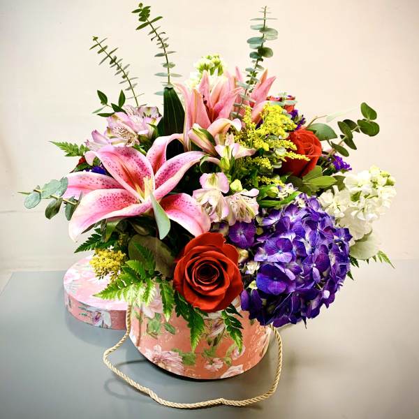 Mixed bouquet with pink lilies, red roses, purple hydrangea, and white blooms in a floral hatbox