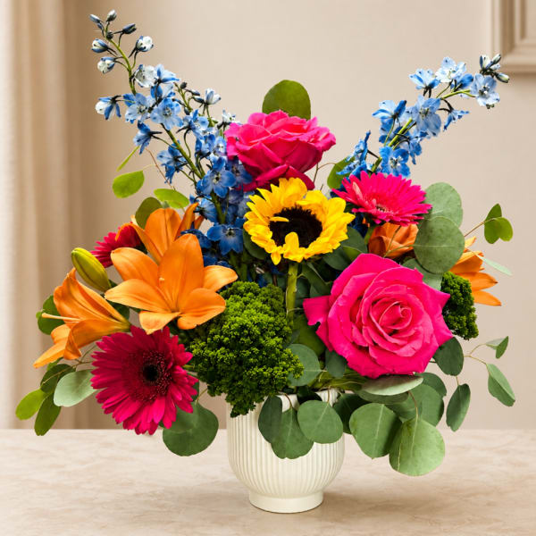 Bright mixed bouquet in a white vase with pink roses, orange lilies, and blue flowers