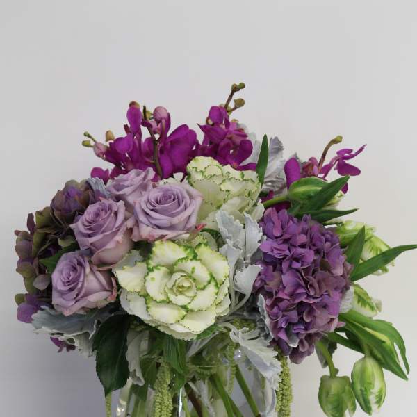 Lavender roses, purple hydrangea, orchids, and green-white blooms in a clear glass vase