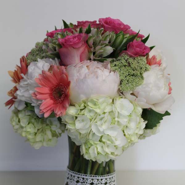 Round bouquet of pink roses, white hydrangeas, and coral gerberas in a glass vase with lace trim